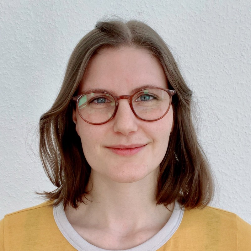 Man sieht eine weiblich gelesene Person im Portraitformat vor grauem Hintergrund. Sie trägt einen senffarbenes T-shirt und eine Brille mit runden Gläsern. Sie lächelt.   