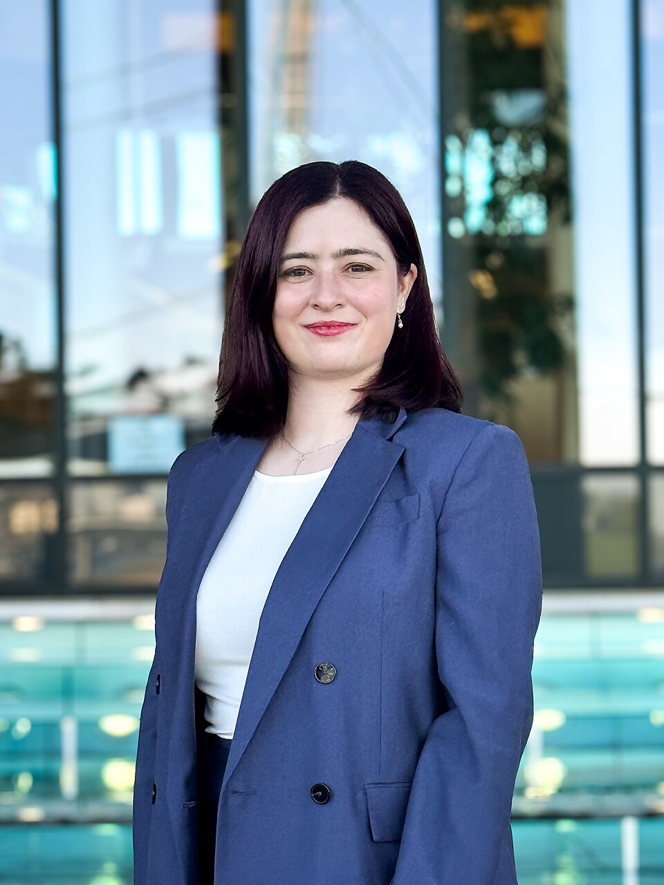 Julia Eisentraut schaut in die Kamera. Sie hat dunkelrote Haare und trägt einen blauen Blazer mit weißem T-Shirt. Im Hintergrund ist verschwommen das Wasserspiel am Landtag zu sehen. 