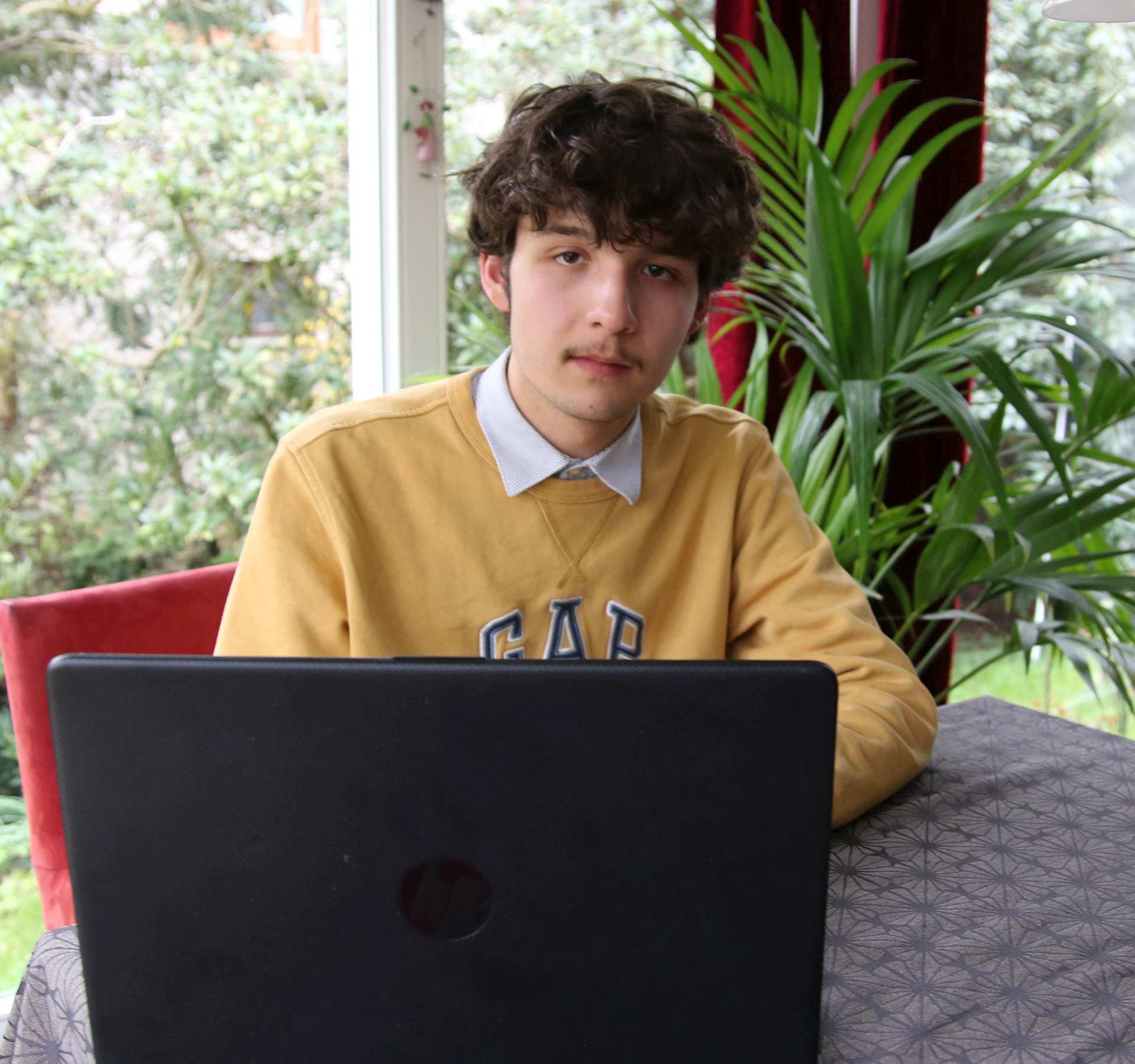 Junger Mann mit gelbem Sweater und Hemdkragen, vor einem Laptop an einem Tisch sitzend
