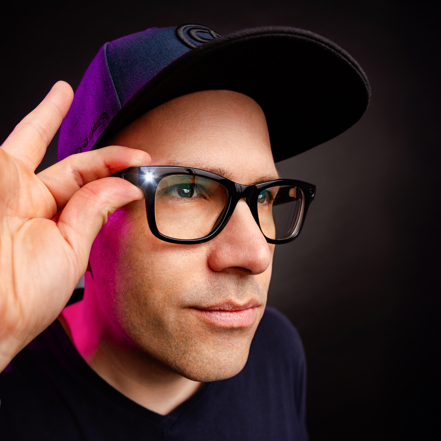 Ein Mann (Thomas Riedel) vor dunklem Hintergrund, ausgeleuchtet mit violetten Farbakzenten von der Seite. Er trägt ein dunkelblaues Sakko, ein graues T-Shirt und eine blaue Basecap mit Logo. Er schaut freundlich in die Kamera und tippt sich mit dem rechten Zeigefinger an die Schläfe bzw. den Bügel seiner schwarzen Brille. Auf dem linken Brillenglas (vom Betrachter aus rechts) ist ein künstlicher, sternförmiger Lichtblitz (Lens Flare) zu sehen, der die Brille als ‚Smart Glasses‘ oder High-Tech-Objekt inszeni