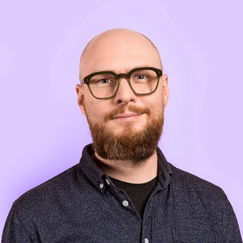Porträtfoto des Speakers: Mann mit Glatze, dunkler eckiger Brille und Vollbart, leicht lächelnd, vor hellviolettem Hintergrund.