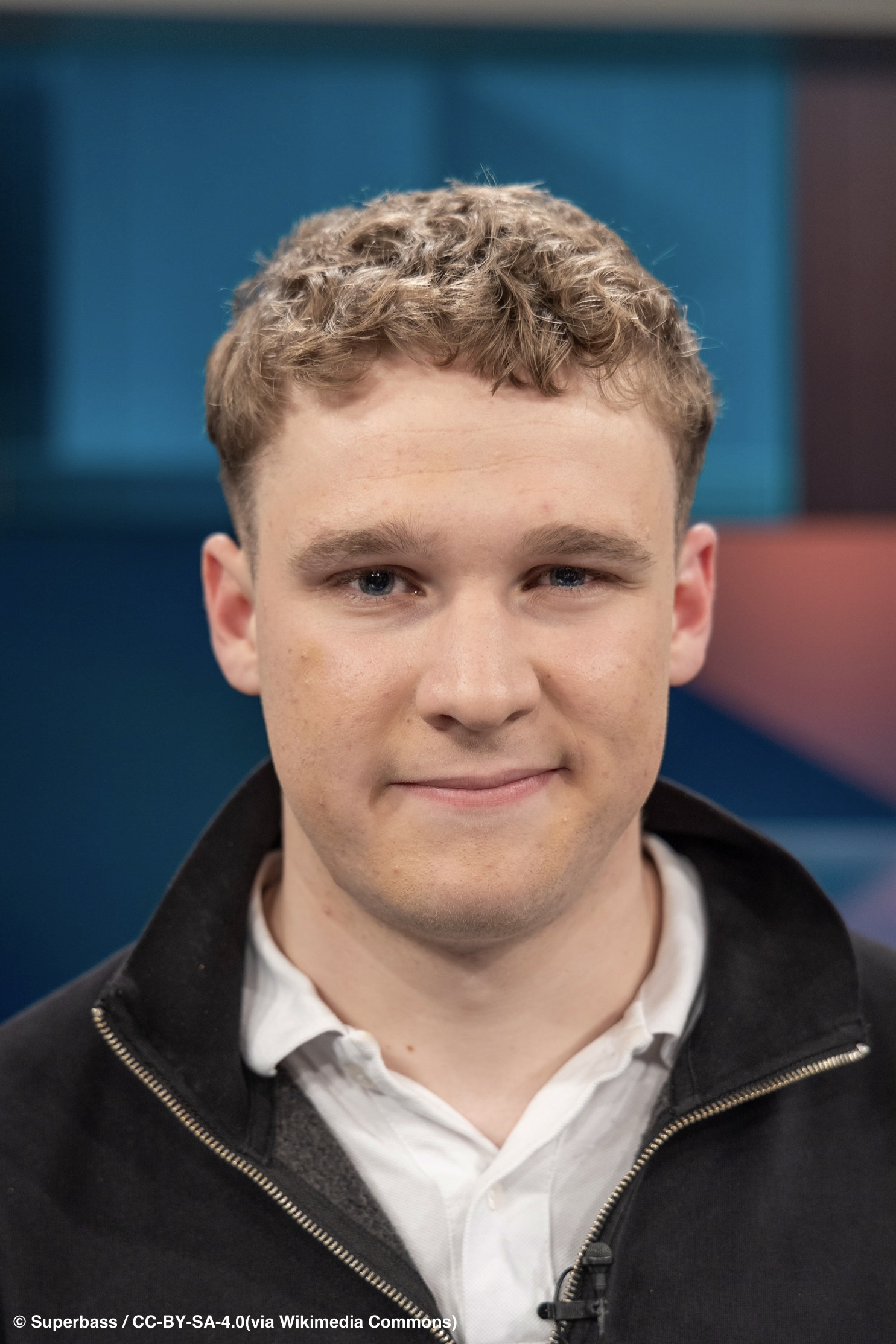 Porträt eines jungen Mannes mit kurzen, lockigen hellbraunen Haaren, weißem Hemd und dunklem Jumper. Er blickt frontal in die Kamera und lächelt leicht. Im Hintergrund sind abstrakte blaue und rötliche Flächen zu sehen.