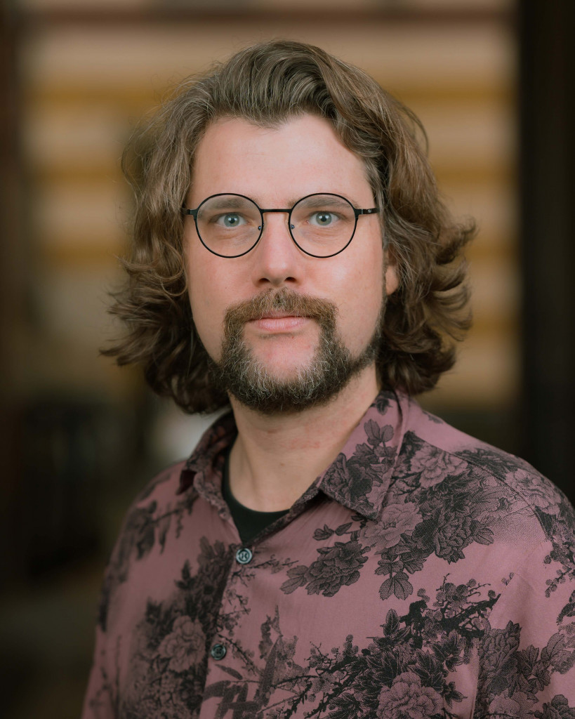 cis-Mann, bärtig, langes Haar, runde Brille, gemustertes Hemd